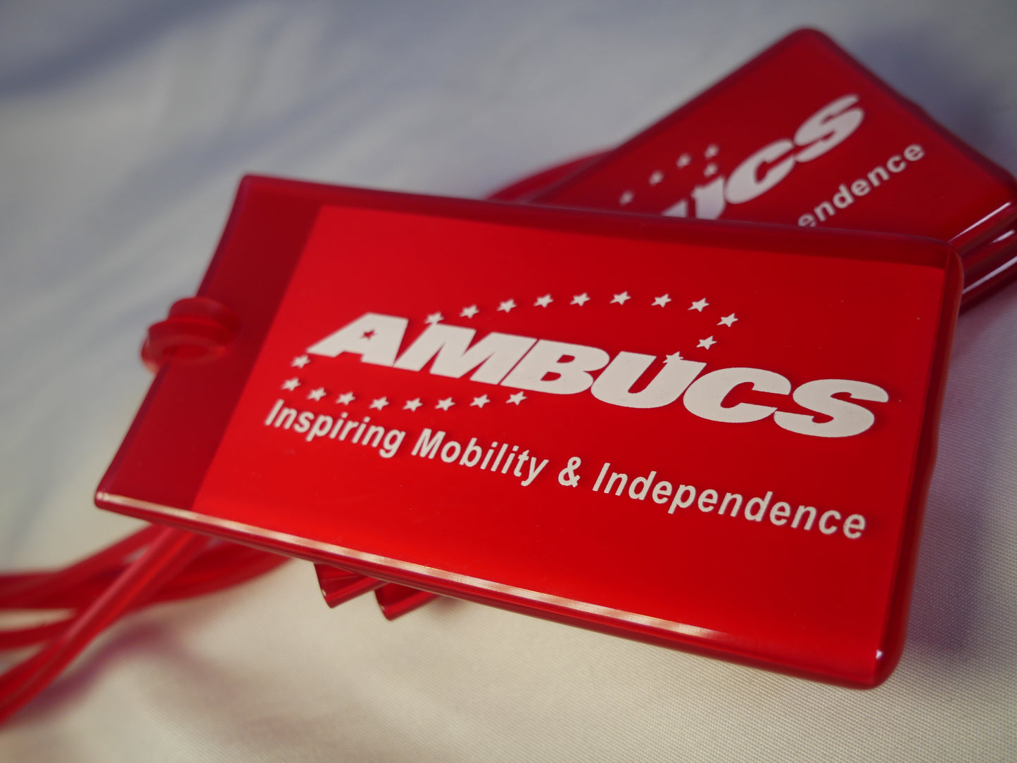 Pictured is a red luggage tag made of soft pliable vinyl with the AMBUCS logo and the words Inspiring Mobility and Independence written in white text.