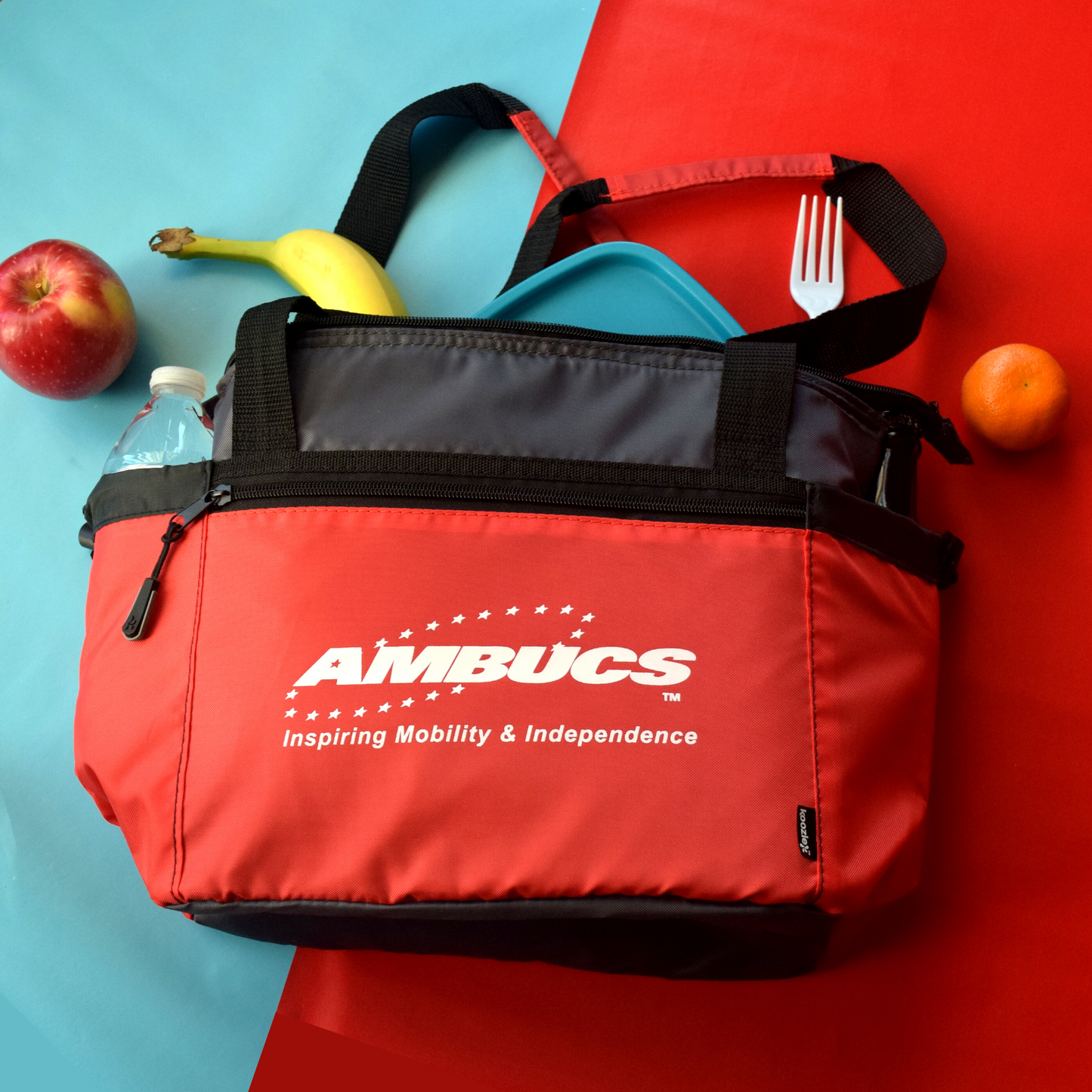 Red insulated zipper cooler bag with black handles. Printed on the front is the AMBUCS logo in white and the words inspiring mobility and independence in white lettering